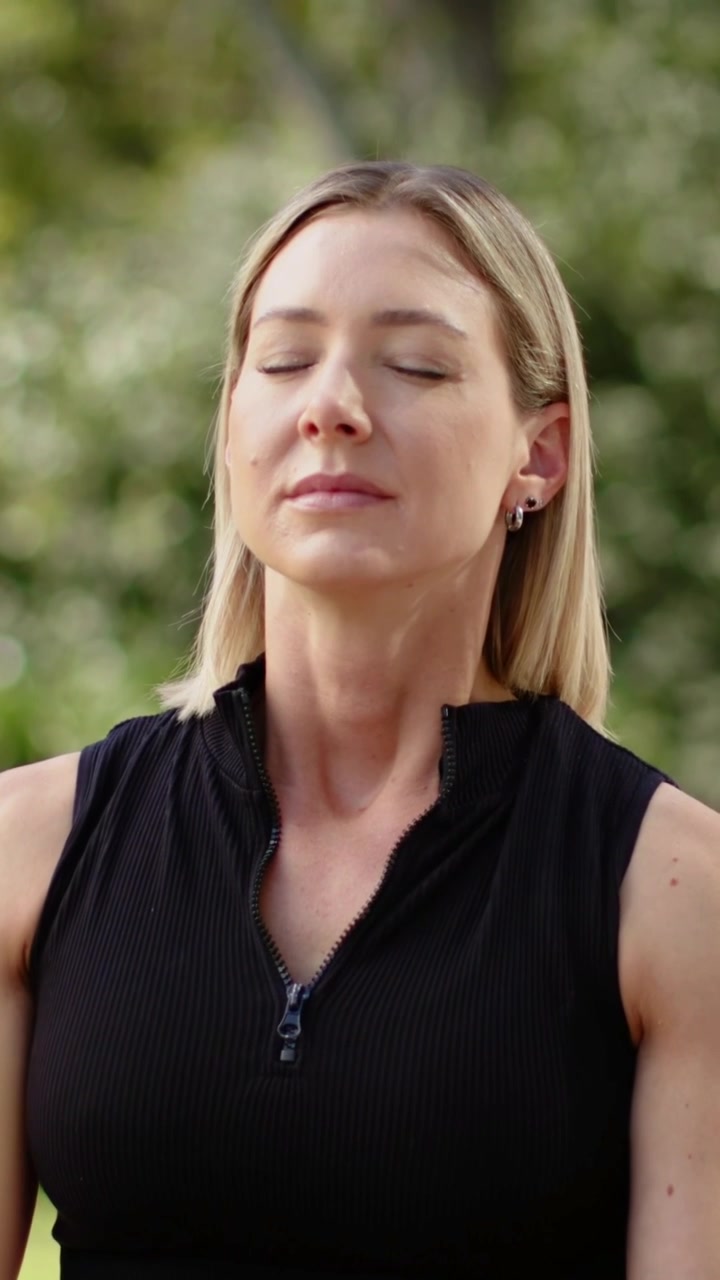 Vídeo vertical: Mudança de foco revelando uma mulher respirando para se acalmar no parque, vestindo um top preto com zíper.