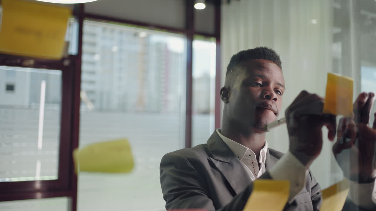 A young serious black business man in a suit creates a board of ideas on a glass wall gluing sticks and signing them.