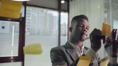a young serious black business man in a suit creates a board of ideas on a glass wall gluing sticks and signing them.