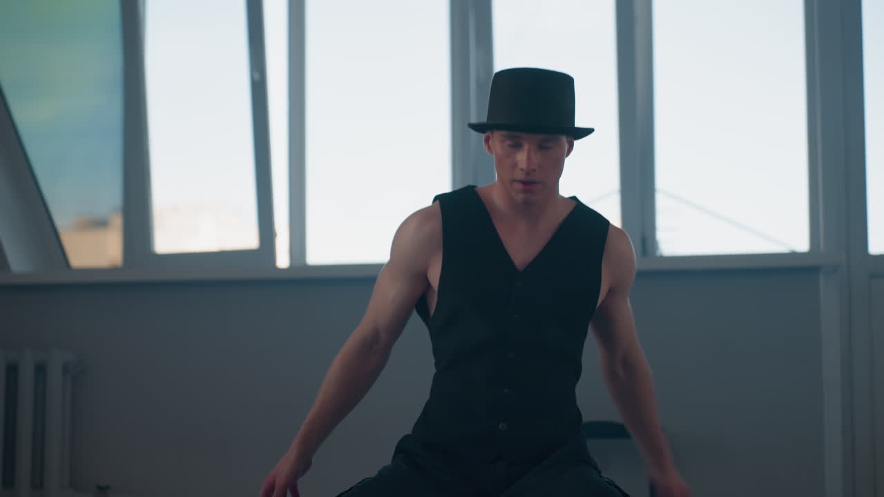 young male dancer kneels on loft studio floor performing expressive moves in black vest and pants with chair silhouette behind windows casting light and smoke adding dramatic urban vibe
