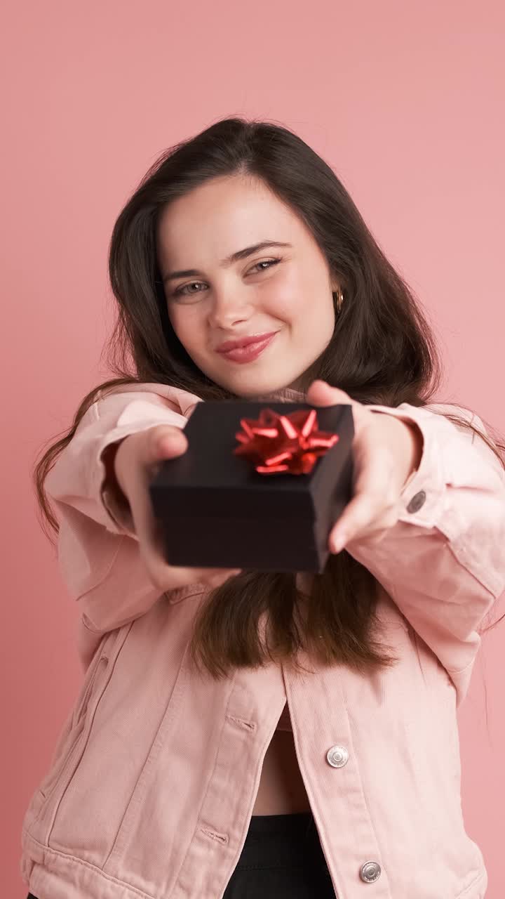Happy woman giving gift box and looking at camera in pink studio