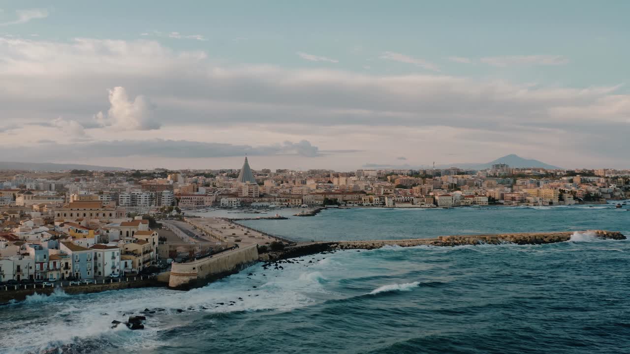 Drone shot above Ortigia island with Mount Etna in a distance. Aerial view over historic buildings on a natural fortress by Mediterranean sea with waves crashing on the coast. UNESCO. Sicily. Italy.