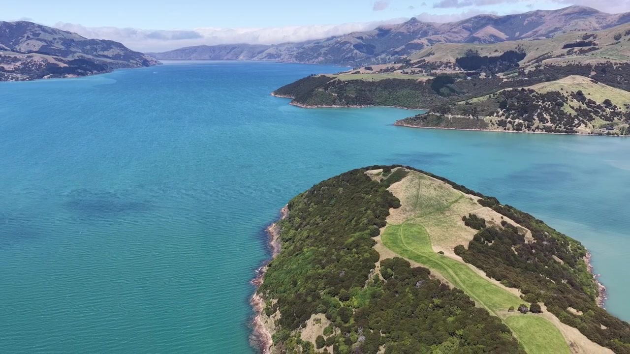 Il drone scende e si allontana sopra una penisola boscosa circondata dalle acque turchesi del porto di Akaroa
