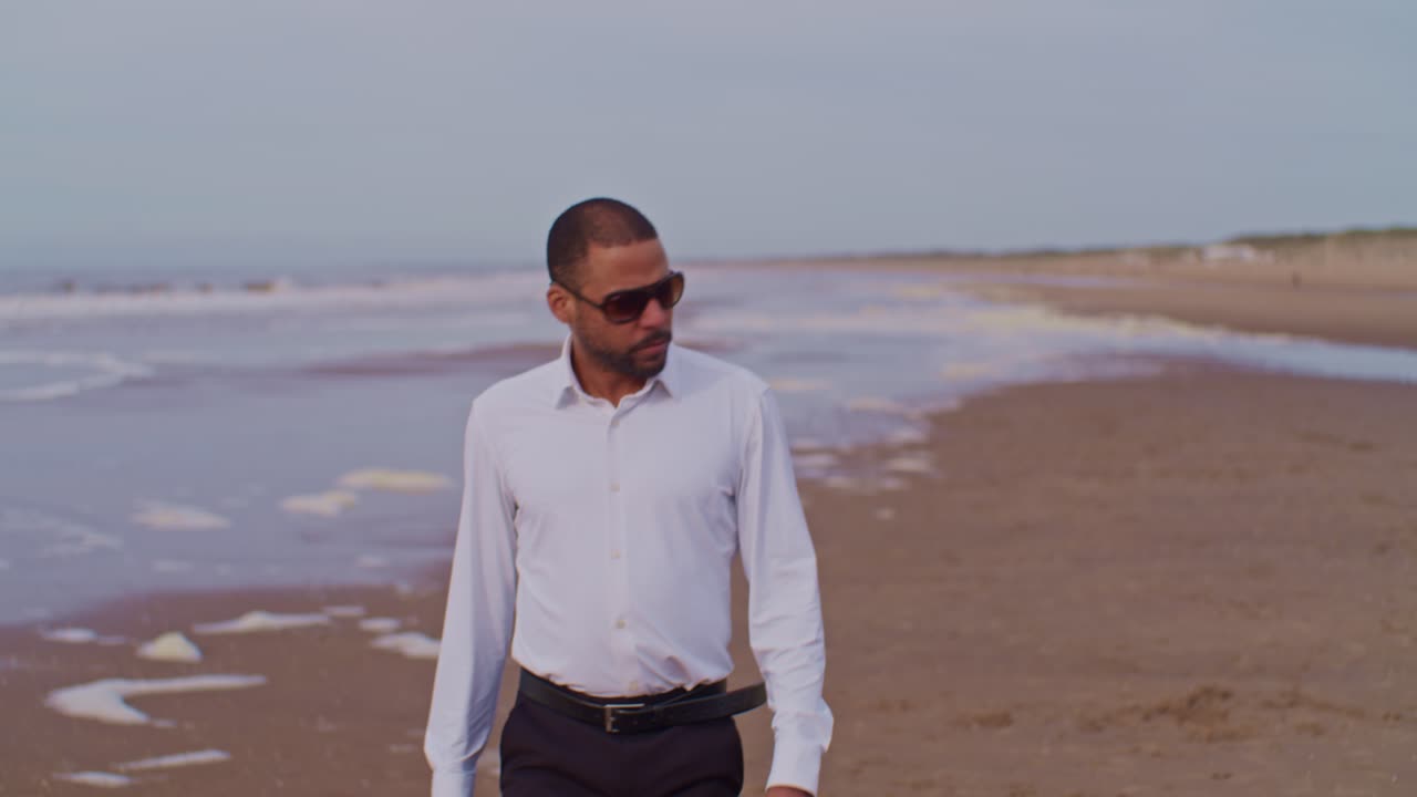 Un modello uomo latinoamericano di colore in abito e camicia bianca cammina sulla riva del mare con occhiali da sole nei Paesi Bassi, L'Aia.
