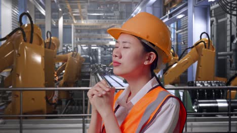 close up side view of asian female engineer with safety helmet pray for something while standing with industrial robotic arms working