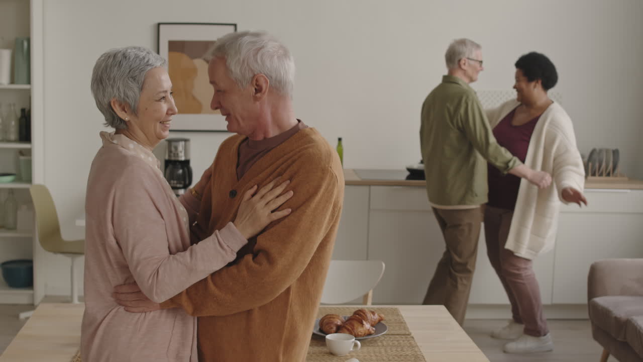 Medium shot of joyful Asian woman talking and dancing with her senior Caucasian husband at home, couple of their friends smiling, moving along on background
