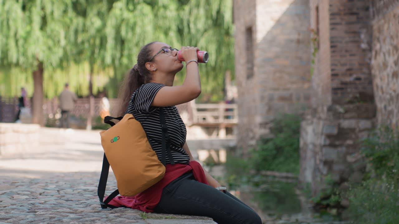 Momento di relax accanto a un vicolo storico. Viaggiatore informale si riposa con una bevanda fresca vicino a un'antica cinta muraria. Turista in abbigliamento casual si ferma a riflettere accanto a una vivace e antica strada acciottolata