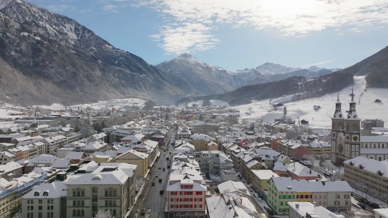 Drone flight over a snow-covered town in the Swiss Alps on a clear winter day. The aerial view reveals the city surrounded by majestic mountains under bright sunlight