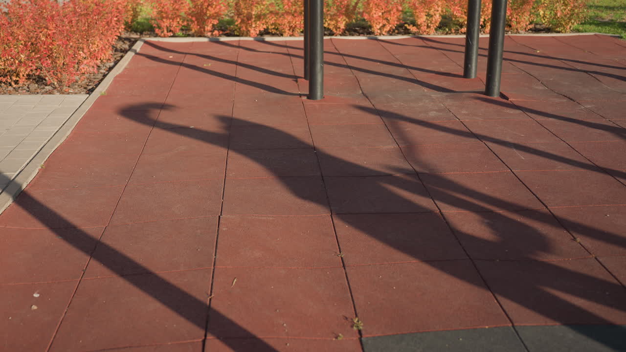 Shadow of workout junkie performing side lunge exercise on outdoor gym court under afternoon sun, long shadows stretching across textured rubber tiles framed by autumn shrubs and metal bars