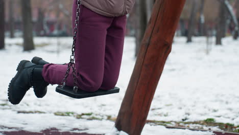 lower body view of person wearing maroon trousers and black boots on swing, with snow-covered ground and blurred background featuring bare trees, building, and passing car