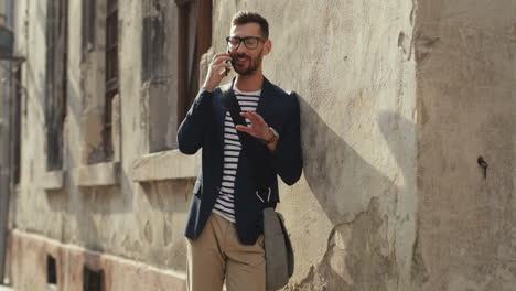 Hombre-Feliz-Y-Elegante-Apoyado-Contra-La-Pared-En-La-Calle-Del-Casco-Antiguo-Y-Hablando-Por-Teléfono-Móvil