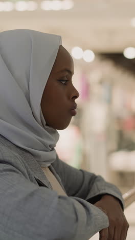 sad african-american woman in hijab leans on transition level handrail in shopping mall closeup. thoughtful muslim lady spends time against blurry shops in mall
