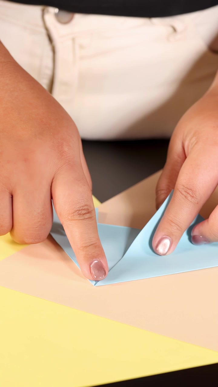 Primo piano di una persona che piega con cura carta origami blu su un tavolo color pastello sotto un'illuminazione morbida e uniforme.