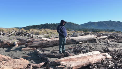 Alter-Mann-Mit-Blauem-Kapuzenpullover,-Der-Im-Sommer-Am-Flussufer-Mit-Totholz-In-Gold-Beach,-Oregon,-Steht