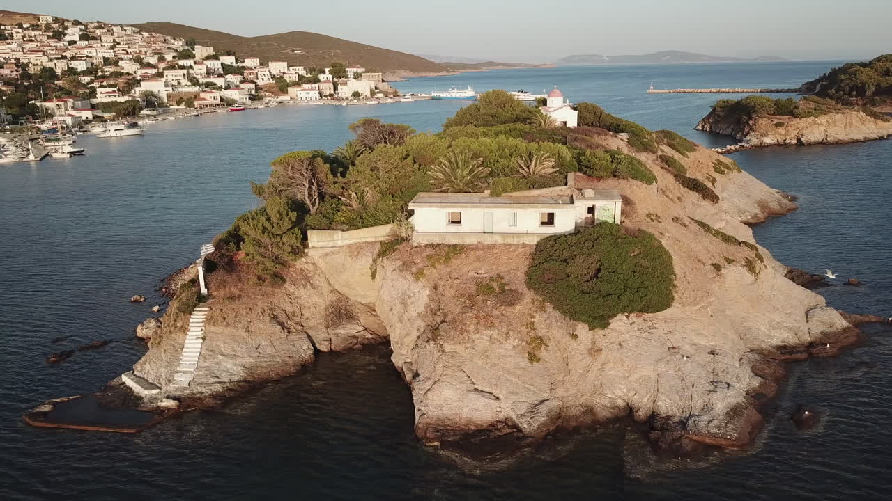 Greece, Ionusses Island, Drone footage turns around the small island with a chapel on the top in front of the bigger island. Behind the small island lays the town center of Ionusses on a sunny day.