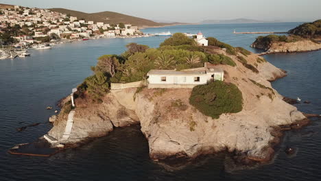 greece, ionusses island, drone footage turns around the small island with a chapel on the top in front of the bigger island. behind the small island lays the town center of ionusses on a sunny day.