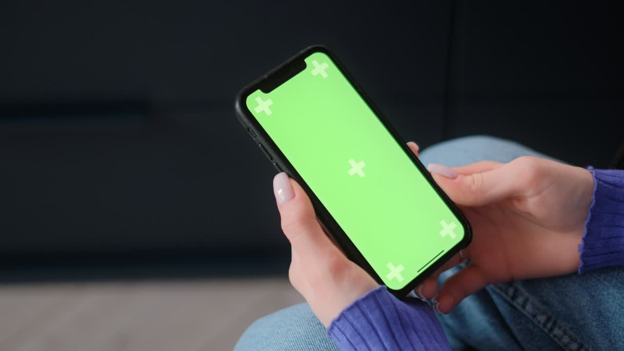 Giovane donna seduta sul divano a casa che usa lo smartphone con schermo verde mock-up. Lo tiene in mano e guarda lo schermo. 4K