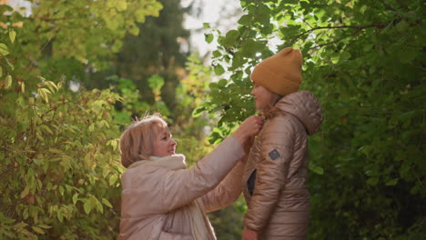 lovely moment with mother gently playing with daughter hair in sunlit autumn woods, warm light filtering through colourful foliage, both in jackets, expressions joyful and tender