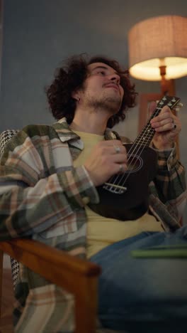 Vertical-close-up-video-of-a-happy-brunette-guy-with-curly-hair-and-a-mustache-in-a-checkered-shirt-playing-a-black-ukulele-near-a-lamp-during-an-atmospheric-evening