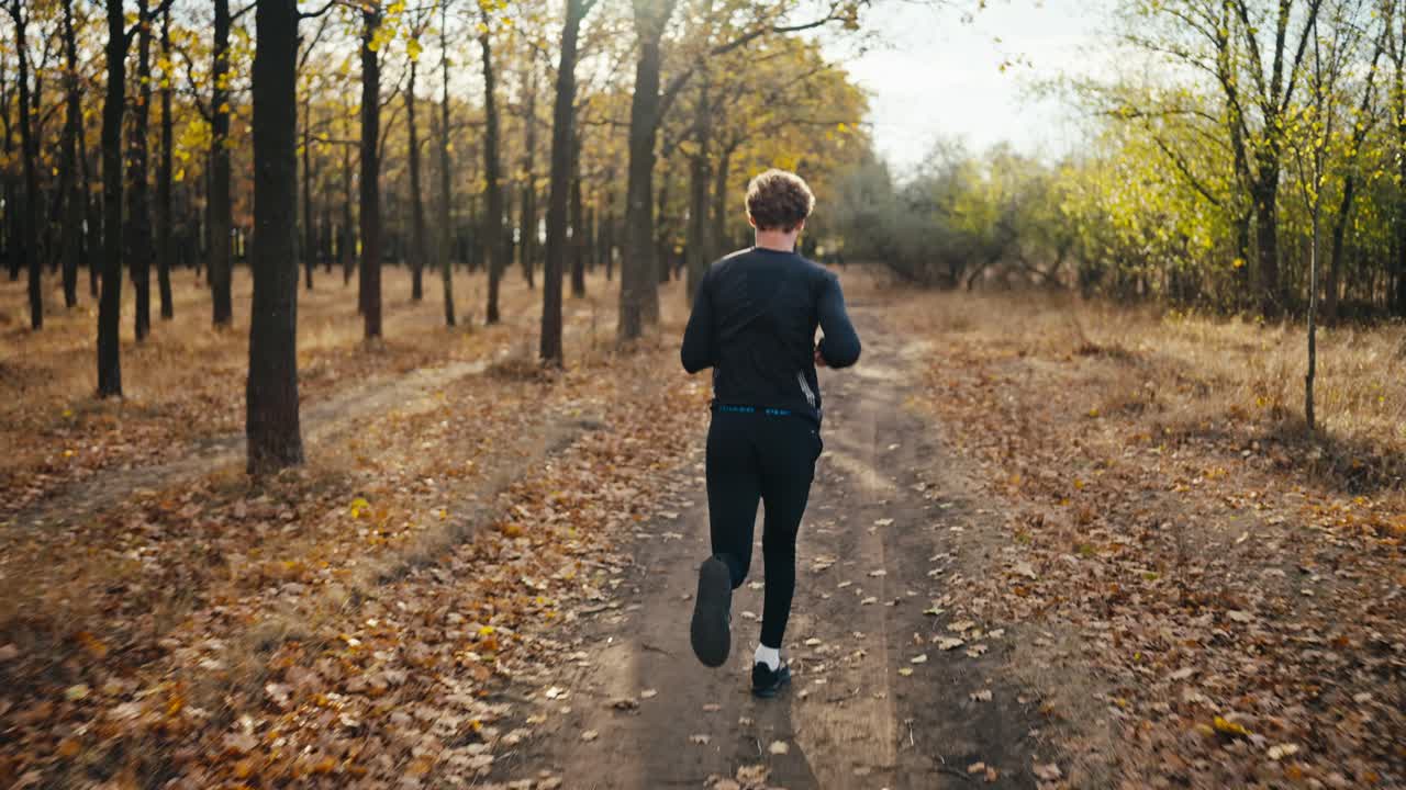 Rear view of a brunette man with curly hair in a black sports uniform runs along an earthen path and looks back at his watch while jogging in the morning in an autumn forest with fallen leaves