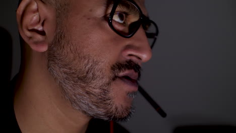 uk asian male wearing glasses conversing using a headset microphone, in dark room, illustrating the concept of telecommuting and remote work