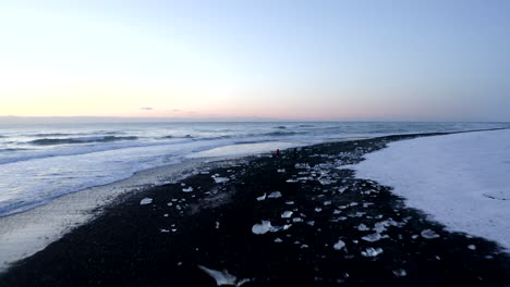 AERIAL:-Flying-over-Black-Beach-with-white-arctic-snow-mountains-in-background-in-Iceland-in-Winter-Snow,-Ice,-Waves,-Water