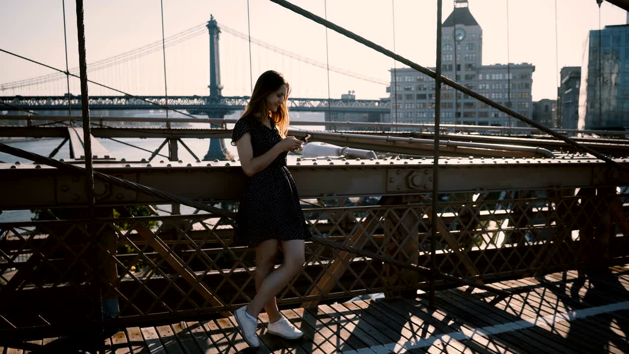 joven chica europea bonita mirando a su alrededor, usando una aplicación de teléfono inteligente en el puente de brooklyn, nueva york increíble vista de la orilla del río 4k