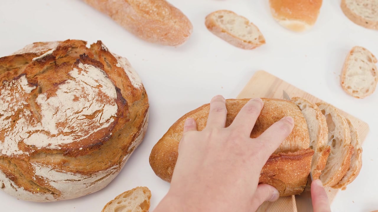 Overhead shot of hands using a knife to slice crusty sourdough bread on white background.