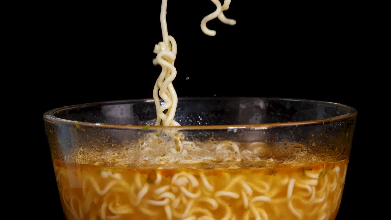 A hand uses chopsticks to lift cooked instant noodles from a glass bowl filled with broth, set against a black background with even lighting and a static camera