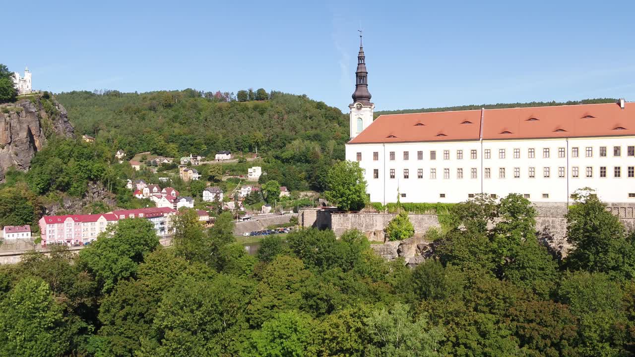 A drone ascends revealing the morning sun hitting residential buildings and the iconic Decin Castle in the background