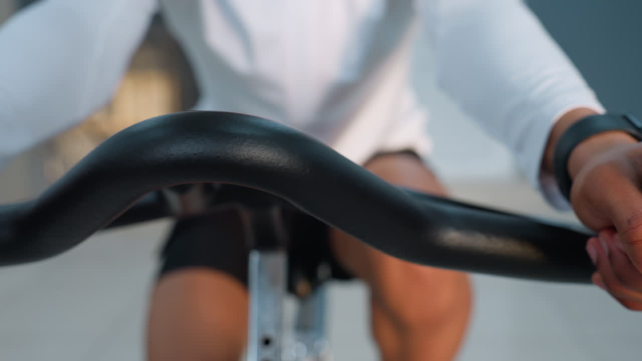 Close up of athlete gripping bike handle bar wearing smart watch during intense indoor cycling workout in modern gym with blurred exercisers and equipment in background