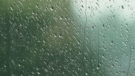 Gotas-De-Lluvia-Deslizándose-Lentamente-Sobre-El-Cristal-De-La-Ventana-En-Un-Día-Lluvioso,-árbol-Verde-Desenfocado-En-El-Fondo,-Toma-De-Primer-Plano-Medio