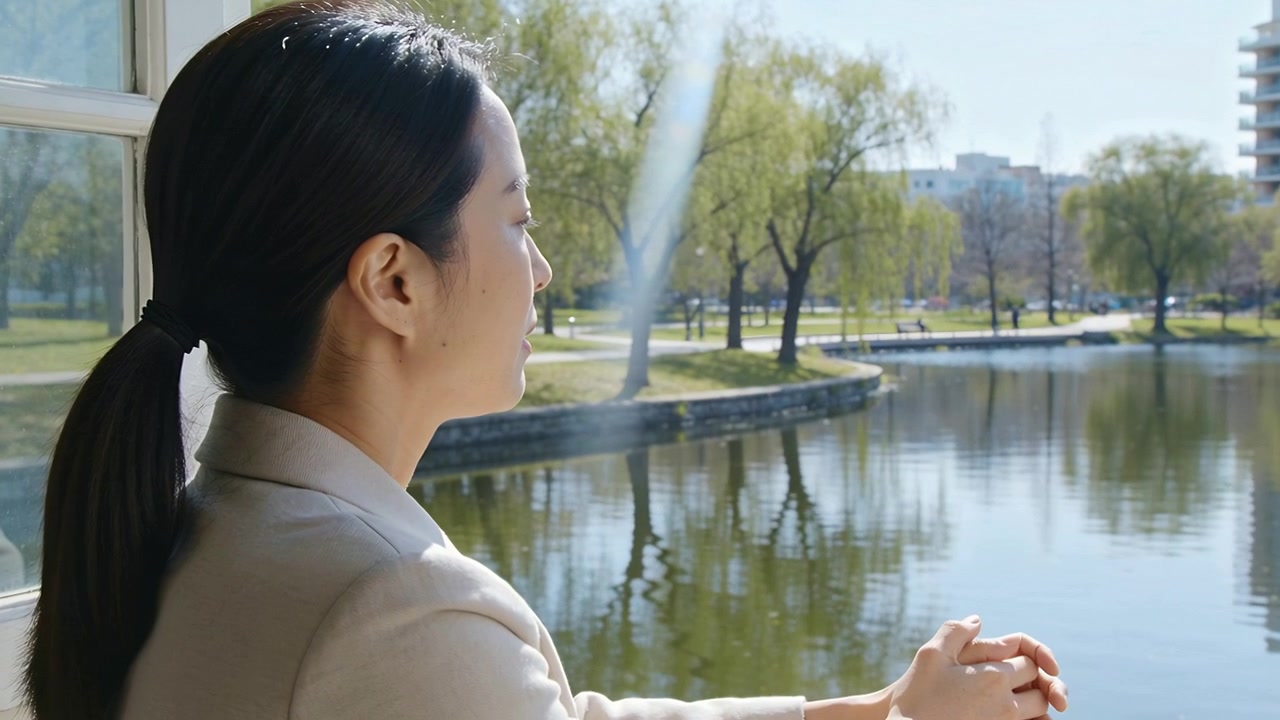 一名穿着米色西装外套的女子睁开眼睛，在阳台栏杆旁沉思并做手势，背景有池塘，可用于添加文字。