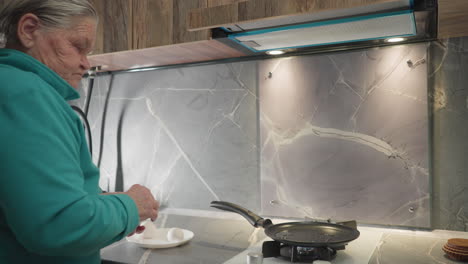 older woman cooking in modern kitchen, adding food to frying pan on stove, with kitchen counter, backsplash, and lighting, preparing meal, focused on senior cooking process and kitchen environment