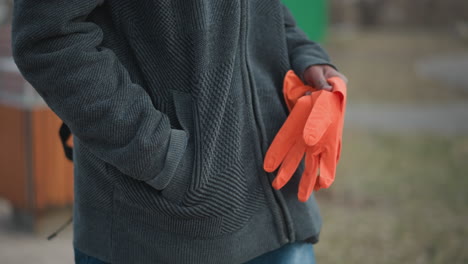 black guy wearing dark gray textured hoodie holding bright orange gloves in one hand with other hand in pocket, standing outdoors on paved path during early spring, blurred park background in natural daylight