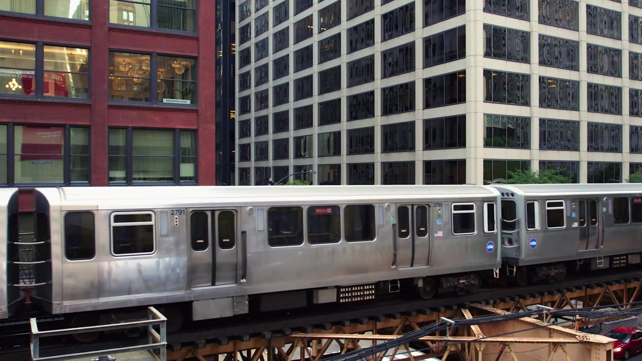 Vista ravvicinata con drone di un treno CTA argentato che viaggia su binari sopraelevati attraverso il centro di Chicago, con edifici per uffici e architettura urbana alle spalle.