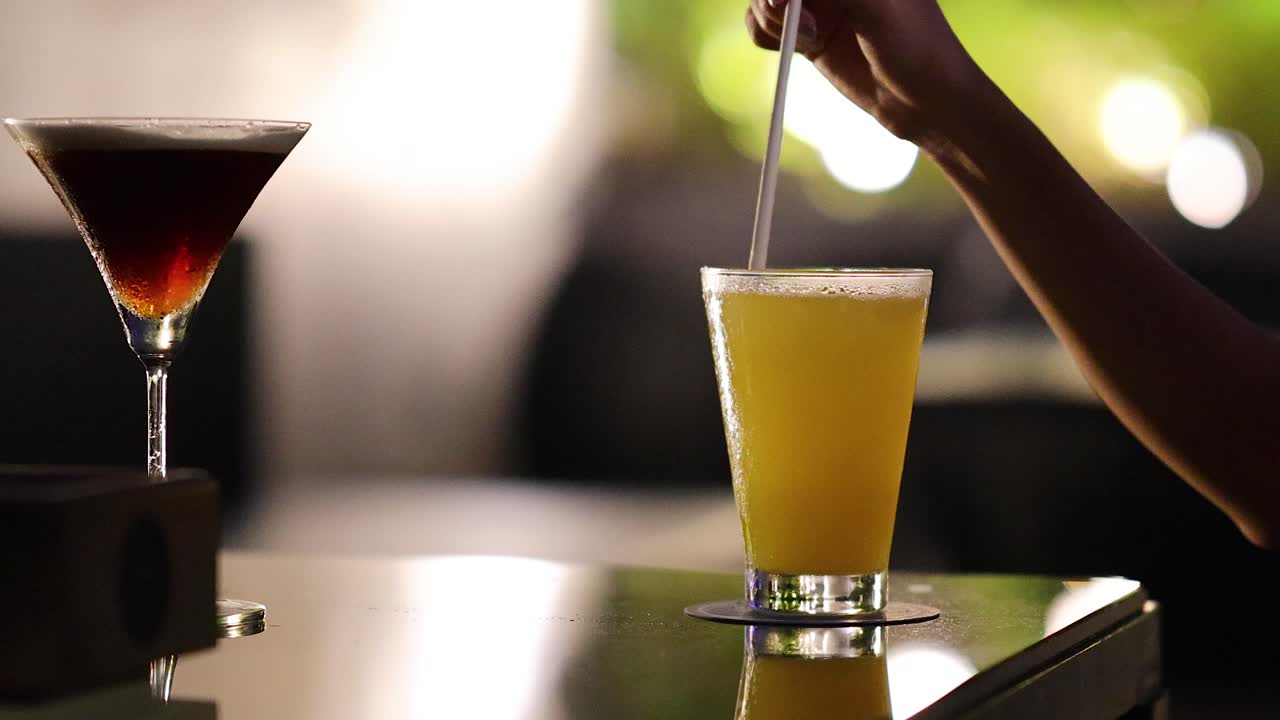 Hand stirs vibrant yellow cocktail beside dark drink on reflective bar in soft evening light