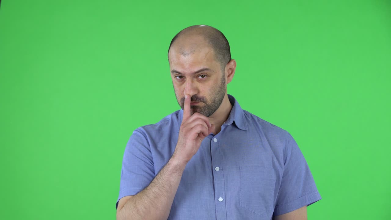 retrato de un hombre de mediana edad mirando a su alrededor y haciendo un gesto de silencio secreto. hombre calvo con barba en camisa azul posando en pantalla verde en el estudio. cerca