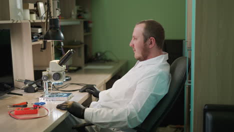 a close view of a technician in a white lab coat relaxing in his seat while carefully examining a circuit under a microscope in his laboratory, he looks up and taps his hand on the table