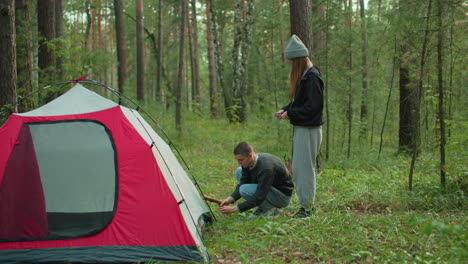 man squats beside tent holding wooden stick while woman stands beside handing him metal peg as they work together to secure tent in forest surrounded by trees and lush greenery