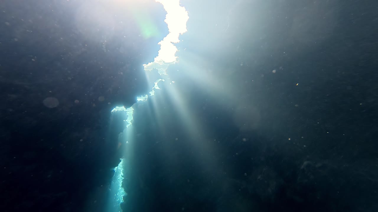 Eksplorasi gua bawah air biru tua dengan sorotan sinar matahari yang menembus formasi batuan bergerigi gelap ke kedalaman laut yang jernih pada hari musim panas yang cerah, dilihat dari sudut kamera mendongak.