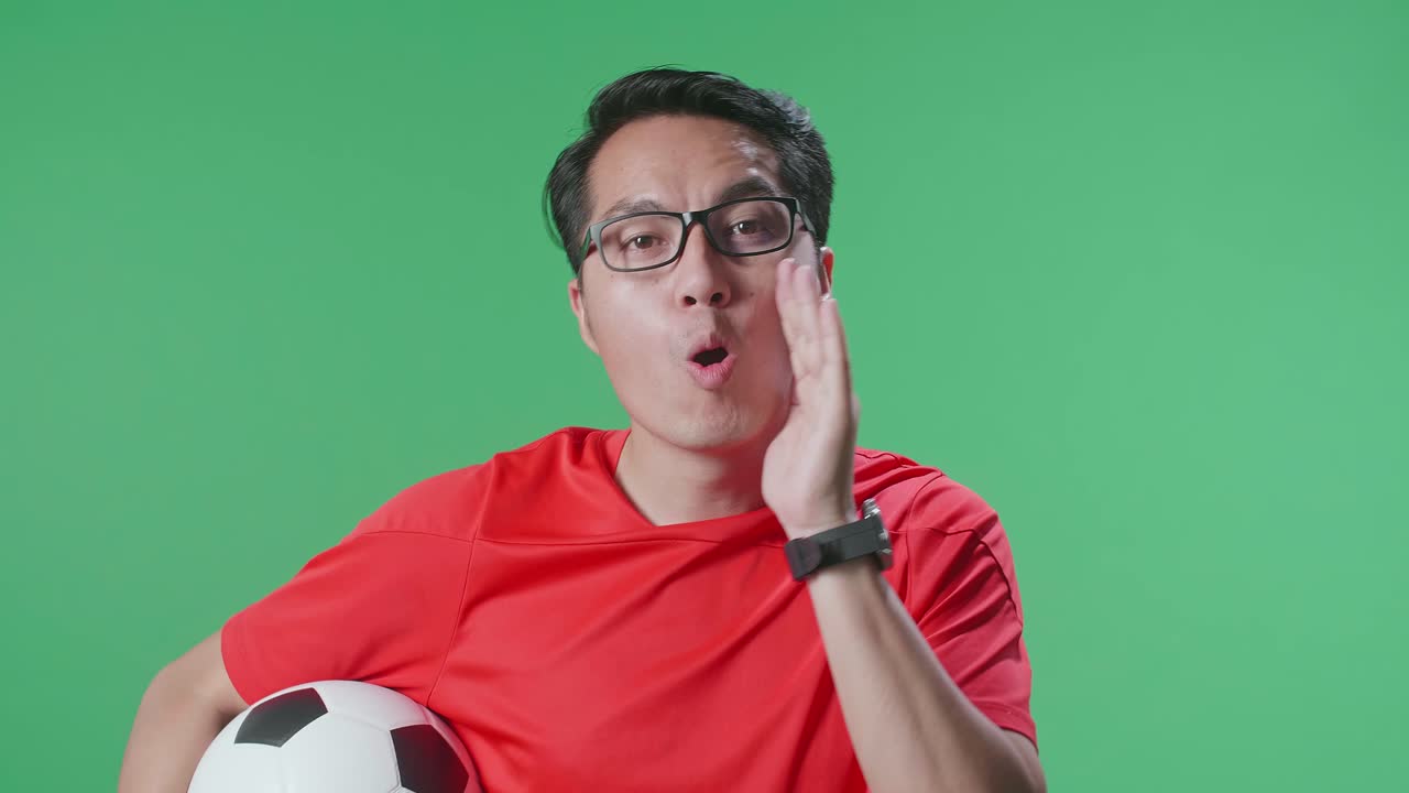 초록색 스크린 배경에서 축구 선수를 응원하는 동안 패자를 조롱하는 공을 가진 아시아 남자의 가까운 모습