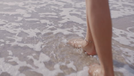 Cerrar-Los-Pies-De-Una-Mujer-Caminando-Descalzo-En-La-Playa-Disfrutando-De-Las-Olas-Salpicando-Suavemente-A-Una-Turista-En-Vacaciones-De-Verano