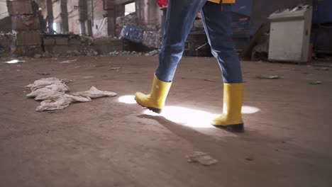 Unrecognizable-woman-in-protective-working-clothes----rubber-gloves-and-boots-walking-by-waste-recycling-factory,-huge-area-with-used-bottles,-recycling-plant