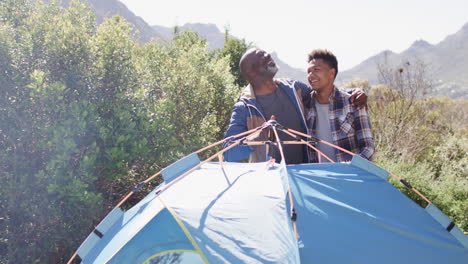 happy african american father and son pitching tent embracing in sunny countryside, slow motion