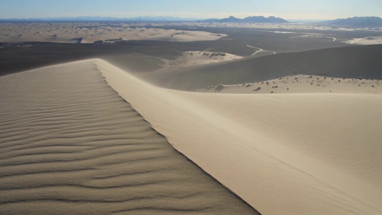 Telecamera drone in movimento che rivela la cresta di una duna di sabbia sopra il deserto, mostrando increspature, la parete sottovento e una catena montuosa distante.