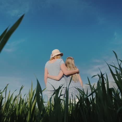 Low-Angle-Shot:-Mom-Hugs-Daughter-Standing-In-A-Beautiful-Green-Meadow-Against-A-Clear-Blue-Sky