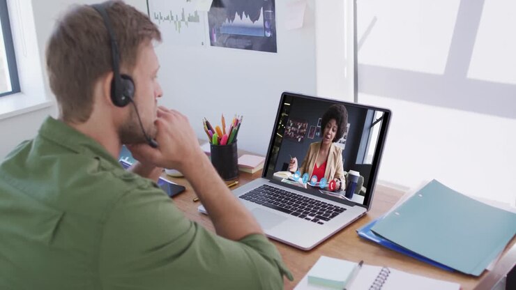 Kaukasisk mand bruger bærbar computer og telefonheadset på videoopkald med en kvindelig kollega.