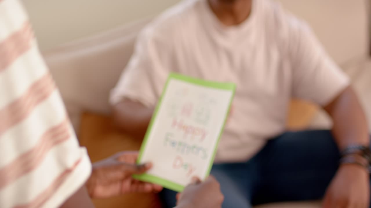 Padre e figlio afroamericani sul divano, il figlio presenta un biglietto fai da te per la Festa del Papà, il padre ride.