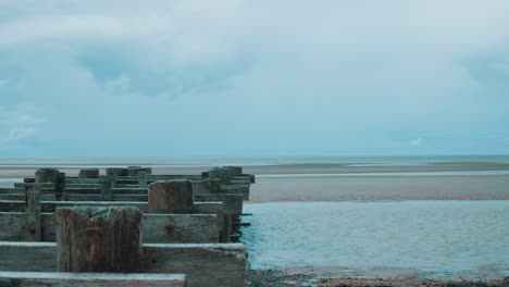 Eine-Kleine-Holzstruktur-Erstreckt-Sich-An-Kleinen-Wasseradern-Vorbei-Und-Sitzt-Bei-Ebbe-Am-Langsam-Trocknenden-Strand,-Während-Sich-Am-Horizont-Ein-Wütender-Sturm-Zusammenbraut